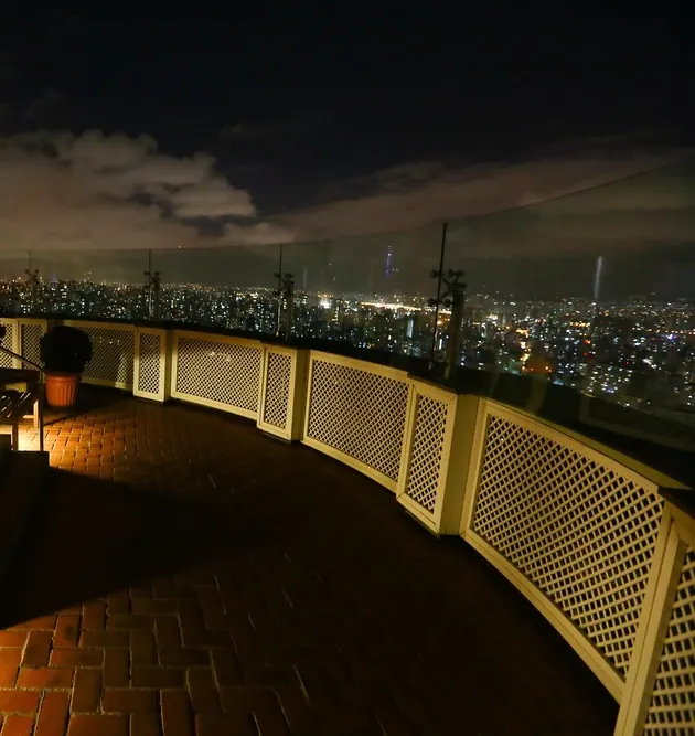 O terraço do mirante à noite, com o piso de tijolos, o parapeito em treliça e a cidade iluminada